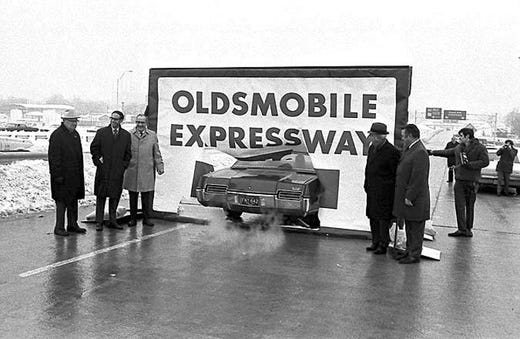 Car driving through a large sign for opening ceremonies of Oldsmobile Expressway with onlookers.